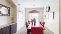 This single-floor space is a refined meeting or private dining room designed with a classic yet bold aesthetic. The centerpiece is a long, rich hardwood table surrounded by tufted red leather chairs, creating a striking visual contrast against the room's warm cream-toned walls and neutral carpet flooring. Natural light filters in through multiple windows, including a distinctive arched window adorned with a red patterned valance at the far end of the room, as well as horizontal blinds along the side wall that offer adjustable light control. A charming circular porthole-style window adds an architectural accent to one wall. Decorative elements include framed artwork and a large wall clock, lending the space a polished, curated character. An arched alcove along one wall reveals a sideboard with a granite countertop and a decorative triple-circle mirror, serving as a functional and stylish accent feature. Recessed lighting in the ceiling provides consistent illumination throughout. A glass-paneled door connects the room to adjacent spaces, enhancing accessibility and openness. Overall, the space combines functional design with elegant detailing, making it well-suited for meetings, private gatherings, or formal dining occasions.