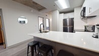 This contemporary urban unit showcases an industrial-modern aesthetic defined by exposed concrete ceilings, light wood-style flooring, and a clean, neutral color palette throughout. The open-concept kitchen serves as a central hub, featuring a large two-tone island with a white quartz countertop, an integrated sink with a matte black faucet, and bar seating for casual dining. White upper cabinetry, stainless steel appliances—including a refrigerator, microwave, and range—and a subway tile backsplash complete the well-equipped kitchen. The living area offers a comfortable arrangement with dark leather seating, a media console, and access to additional spaces, all beneath the striking exposed concrete ceiling. Multiple bedrooms are thoughtfully designed, each accommodating a bed with under-bed storage, a dedicated desk and shelving unit, and natural light from sizeable windows that frame urban views. The bathroom area is split into a shared vanity zone with dual sinks, gray cabinetry, and tiled accents, and a separate wet room with a full soaking tub and shower combination. Wood-look flooring flows consistently throughout, tying the industrial and contemporary design elements together into a cohesive and functional living environment.