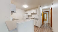 This single-floor residence offers a well-organized layout designed for comfortable everyday living. The kitchen is a standout feature, designed in a galley style with crisp white cabinetry, brushed nickel hardware, light wood-look flooring, and a full suite of white appliances including a range, refrigerator, and dishwasher. A peninsula counter with additional storage extends the kitchen workspace and creates a natural division between the cooking area and the rest of the home. The spacious living area is bathed in natural light from a large sliding glass door that opens to an outdoor space, complemented by light wood-look flooring and a textured ceiling. Baseboard heating runs along the walls, ensuring warmth throughout. The bedrooms are generously sized with soft carpeting underfoot, each featuring windows that frame lush green outdoor views. At least one bedroom includes a walk-in closet with built-in shelving for ample storage. The bathroom is clean and functional, featuring a full soaking tub with an overhead shower, white surround panels, and wood-look flooring. Throughout the space, a neutral color palette of whites and warm beiges creates a bright, cohesive, and move-in-ready atmosphere.