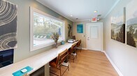 Located at 2870 Regal Cir in Hoover, Alabama, this single-room cyber café office is a thoughtfully designed shared workspace that maximizes functionality within a compact footprint. The room features light hardwood flooring, sage-green accent walls, and crisp white trim with crown molding, creating a clean and welcoming aesthetic throughout.

A long, continuous white countertop workstation runs along one wall, accompanied by multiple wooden chairs that provide ample seating for several users simultaneously. The workstation is equipped with a desktop computer, keyboard, printer, and free Wi-Fi access, making it a practical hub for residents' everyday computing needs.

A large picture window dominates one wall, offering an expansive view of the outdoor pool area and surrounding wooded landscape, filling the space with natural light. Recessed ceiling lighting supplements the natural illumination. Decorative artwork, including landscape paintings and a bold abstract canvas, adds character to the walls.

An adjacent doorway connects to a fitness center, broadening the utility of the space within the larger community amenity area. Informational signage and cyber café usage guidelines are clearly posted, reflecting an organized and community-focused environment.