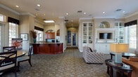 This thoughtfully designed single-floor community space offers a warm and inviting atmosphere, combining functional amenities with refined interior details. The layout flows seamlessly between distinct zones, each complementing the next with consistent design elements including crown molding, recessed lighting, and a neutral warm color palette throughout.

The lounge area serves as the heart of the space, featuring a built-in white display cabinet with glass doors flanking a wall-mounted television, elegant arched architectural accents, and large windows dressed with navy valances that allow generous natural light while framing views of the surrounding landscape. Comfortable seating, including a plush sofa and patterned accent chairs, creates an approachable and relaxed environment.

Adjacent to the lounge, the kitchen area is equipped with rich cherry wood cabinetry, expansive granite countertops, stainless steel appliances, and a light blue tile backsplash that adds a subtle pop of color. A generous peninsula provides additional workspace and serves as a natural gathering point.

The dining area features abundant natural light pouring in through tall windows and glass French doors that open to an outdoor area, offering a bright and airy setting. Round and bistro-style tables provide flexible seating arrangements. Together, these spaces create a cohesive, welcoming environment well-suited for community gatherings and everyday use.