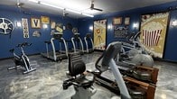 This single-floor fitness room offers a creative and energetic environment for exercise, distinguished by its striking Hollywood/cinema theme. The walls are painted in deep navy blue and adorned with vintage movie-themed decorations, including film reels, ticket stubs, clapperboards, and a bold retro movie camera mural serving as a dramatic focal point. A "Silence Please" sign and vintage clock add to the cinematic atmosphere.

The space is well-equipped with multiple treadmills arranged along one wall, elliptical machines, a recumbent bike, and a rowing machine, providing a comprehensive cardio workout experience. Two ceiling fans ensure proper air circulation throughout the room. The flooring features a polished, marbled epoxy finish in grey and silver tones, adding a sleek, modern contrast to the themed décor.

Wall-mounted lighting fixtures complement the overhead fans, providing adequate illumination for workouts. The overall ambiance balances a fun, movie-inspired aesthetic with a fully functional fitness environment, making it an engaging and motivating space for exercise.
