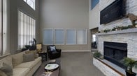 This well-appointed single-floor space makes an immediate impression with its dramatic double-height ceilings that lend an expansive, airy quality to the main lounge area. The generous vertical space is accentuated by soft neutral wall tones in warm gray, complemented by a bold blue accent near the upper portion of the walls, creating a modern yet inviting atmosphere.

The lounge area is thoughtfully furnished with a comfortable sofa and armchair arranged around a dark wood coffee table, offering a relaxed setting for gathering or unwinding. A prominent stacked-stone fireplace anchors one side of the room, topped with a large wall-mounted flat-screen television, making it a natural focal point. The fireplace surround features clean, contemporary stonework that adds texture and warmth to the space.

Four frosted-glass panel windows are mounted along the main wall, allowing diffused light to filter through while maintaining a sense of privacy. Large windows along the side wall welcome additional natural light into the space. Plush carpeting throughout the lounge area enhances the comfortable, welcoming feel.

An adjacent room visible in the background suggests additional functional space, potentially a fitness or activity area. Overall, the space balances sophisticated design with practical comfort, making it ideal for community gathering or professional use.