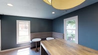 Located at Lake Castleton, this single-floor space presents a thoughtfully designed interior with a contemporary aesthetic. The main lounge area is defined by deep blue-gray walls that create a calm, sophisticated atmosphere. Built-in banquette seating with striped upholstery lines one wall, complemented by small round bistro tables with marble-style tops and slender metal bases — ideal for casual gathering or informal meetings.

A substantial wood-top counter anchors the space, adding warmth and contrast against the cooler wall tones. Overhead, a large drum pendant light with a warm gold shade provides ambient illumination, while recessed lighting supplements the natural light that pours in through multiple white-framed windows. The windows offer views of mature trees and the surrounding landscape, further enhancing the inviting atmosphere.

Tile flooring in a wood-plank pattern flows through the space, lending continuity and a modern touch. An adjacent area features a marble-topped built-in desk surface paired with a contemporary white chair, suggesting a functional workstation. Mechanical and utility areas are also present, housing essential building systems. Overall, the space balances practical functionality with refined, modern design details.