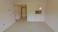 This single-floor space offers a straightforward and functional layout characterized by clean, neutral finishes throughout. The main living area is finished with light-toned wall-to-wall carpeting, creating a quiet and comfortable atmosphere. A notable architectural detail is the pass-through window cut into the wall between the living area and the kitchen, which allows for easy communication and serving between the two spaces. A pendant light fixture hangs above the pass-through, providing focused illumination and a subtle design element against the otherwise minimal décor. The kitchen area, partially visible through the opening, features cabinetry and a sink. Moving toward the back of the space, a hallway corridor transitions from carpet to tile flooring, leading to the unit's front entry door. The corridor is finished with recessed ceiling lighting, contributing to a well-lit passageway. An additional door is visible along the hallway, suggesting access to a closet or secondary room. The overall color palette is composed of soft whites and beiges, lending the space a clean, neutral, and move-in-ready appearance throughout.