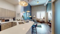 This single-floor residence showcases a compelling blend of industrial and modern design, highlighted by dramatically high ceilings and exposed brick wall details that lend the space a distinct loft-like character. The open-concept living and kitchen area forms the heart of the unit, where hardwood-style flooring, a bold teal accent wall, and ceiling fans overhead create a dynamic yet comfortable atmosphere. A large kitchen island with a white quartz countertop and double undermount sink anchors the space, complemented by light wood cabinetry, a subtle tile backsplash, and stainless steel appliances. Oversized pendant lights hang above the island, adding warmth and visual interest. The living area offers generous space for seating arrangements, with natural light filtering through wide windows dressed with horizontal blinds. The bedrooms provide a more intimate atmosphere, one featuring a deep navy accent wall with carpeted flooring, while another benefits from bright windows and elevated ceilings. The bathrooms are finished consistently with light wood vanities, white quartz countertops, frameless mirrors, and a glass-enclosed walk-in shower with classic subway tile, maintaining a clean, cohesive aesthetic throughout.