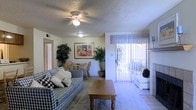 This single-floor residence is thoughtfully arranged to maximize functionality and comfort across its well-defined living spaces. The floor plan reveals a central living and dining area that serves as the heart of the home, anchored by a fireplace that adds warmth and a focal point to the space. Ceiling fans fitted with integrated lighting fixtures are present throughout, contributing to both airflow and ambient illumination beneath textured ceilings.

The living area is open and inviting, with neutral-toned walls and carpeted flooring that transitions to tile in key areas. A sliding glass door framed by vertical blinds and valance curtains draws natural light into the main living space while providing direct access to an outdoor area, extending the sense of openness. A dining nook positioned adjacent to the living room offers a casual seating arrangement.

The kitchen is equipped with standard appliances and cabinetry, with a pass-through or bar area connecting it to the dining space. Multiple bedrooms are situated toward the wings of the floor plan, offering privacy from the main living areas. The overall layout is practical and cohesive, with a straightforward design that prioritizes ease of movement and livability throughout the home.