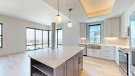 This thoughtfully designed two-bedroom, three-bathroom high-rise residence offers a refined urban living experience with contemporary finishes and sweeping city views. The open-concept kitchen and living area serves as the heart of the home, anchored by a striking powder-blue island topped with white quartz and illuminated by globe pendant lights with brass accents. White shaker cabinetry, stainless steel appliances, and a farmhouse sink complete the kitchen, which flows seamlessly into the spacious living area bathed in natural light from expansive floor-to-ceiling windows. Light hardwood flooring runs throughout the entire residence, lending warmth and continuity. The primary bedroom is generously sized, featuring large windows with panoramic city views and access to well-appointed walk-in closets with customizable built-in shelving systems. The second bedroom is bright and airy, with its own city-facing window. The bathrooms are elegantly finished with white shaker vanities, brass fixtures, large-format tile flooring, and a glass-enclosed tub and shower combination with vertically tiled surround. A clean, bright hallway with recessed lighting connects the private quarters. The overall ambiance is modern, polished, and flooded with natural light.