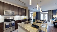 This well-appointed single-floor residence showcases a thoughtfully designed open-concept layout that seamlessly connects the kitchen and dining areas. The kitchen is a standout feature, boasting rich dark-stained cabinetry that provides ample storage alongside a striking granite-topped island with an undermount sink. The island doubles as a breakfast bar, offering casual seating and a natural gathering point for the space. Stainless steel appliances, including a built-in microwave and electric range, complement the subway tile backsplash and add a polished, contemporary feel. Track lighting runs along the ceiling, providing flexible, targeted illumination throughout the kitchen, while elegant cylindrical pendant lights hang above the island for a refined finishing touch. The dining area flows naturally from the kitchen, bathed in natural light streaming through large French doors adorned with draped curtains. A bold navy accent wall anchors the dining space, adding depth and a sophisticated contrast to the lighter tones found throughout. The overall ambiance is modern and inviting, blending functional design with upscale finishes to create a space that is both practical and visually compelling.
