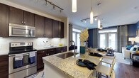 This well-appointed single-floor residence showcases a thoughtfully designed open-concept layout that seamlessly connects the kitchen and dining areas. The kitchen is a standout feature, boasting rich dark-stained cabinetry that provides ample storage alongside a striking granite-topped island with an undermount sink. The island doubles as a breakfast bar, offering casual seating and a natural gathering point for the space. Stainless steel appliances, including a built-in microwave and electric range, complement the subway tile backsplash and add a polished, contemporary feel. Track lighting runs along the ceiling, providing flexible, targeted illumination throughout the kitchen, while elegant cylindrical pendant lights hang above the island for a refined finishing touch. The dining area flows naturally from the kitchen, bathed in natural light streaming through large French doors adorned with draped curtains. A bold navy accent wall anchors the dining space, adding depth and a sophisticated contrast to the lighter tones found throughout. The overall ambiance is modern and inviting, blending functional design with upscale finishes to create a space that is both practical and visually compelling.