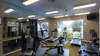 This fitness center is a thoughtfully designed, single-floor exercise facility that provides a comprehensive workout environment. The space is lined with floor-to-ceiling mirrors along one wall, visually expanding the room and allowing users to monitor their form during exercise. Large windows along the opposite wall flood the interior with natural light and offer pleasant views of surrounding greenery, creating an inviting and energizing atmosphere.

The cardio section is well-appointed with multiple treadmills and an upright stationary bike, all positioned to take advantage of the outdoor views. For strength training, the space includes a multi-functional cable and weight machine, along with an adjustable workout bench, catering to a variety of exercise routines. Rubber floor mats are laid beneath the equipment to provide stability and protect the flooring.

Overhead, flush-mounted fluorescent lighting ensures bright, even illumination throughout the facility, while a ceiling fan aids in air circulation during intense workouts. A wall clock keeps exercisers on schedule. The warm, neutral-toned walls and wood-style flooring contribute to a clean and welcoming aesthetic. Overall, this fitness center balances functionality and comfort, making it a practical and pleasant space for a wide range of fitness activities.
