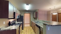 This single-floor space is designed with practicality and comfort in mind. The kitchen serves as a central highlight, featuring a classic galley layout that maximizes efficiency. It is equipped with stainless steel and black appliances, including an electric range, refrigerator, built-in microwave, and dishwasher. The countertops display a granite-look finish that adds a touch of elegance, while the warm-toned wood cabinetry provides ample storage above and below. A double-basin sink is conveniently positioned along one side, complemented by additional counter space ideal for meal preparation. The kitchen also includes a peninsula or pass-through area, offering extra workspace or casual dining potential. Light wood-style flooring runs throughout, lending a clean and cohesive aesthetic to the space. A doorway visible from the kitchen leads to an adjacent room, suggesting a well-connected floor plan. Overhead, a flush-mount light fixture provides bright, even illumination, and the textured ceiling adds a subtle architectural detail. Overall, the space offers a well-organized, functional layout suited for everyday living.