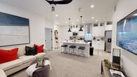 This well-appointed single-story residence showcases a thoughtfully designed open-concept floor plan that seamlessly connects the living and kitchen areas. The kitchen is a standout feature, outfitted with crisp white shaker cabinetry reaching to the ceiling, glass-front upper cabinets, a white quartz countertop island with an undermount sink and black matte faucet, and a suite of stainless steel appliances including a built-in microwave, range, and refrigerator. A sleek stainless steel range hood serves as a focal point above the cooktop, while black pendant lights add a modern contrast. Light wood-tone flooring runs through the kitchen, transitioning to plush carpet in the living area, where a ceiling fan provides comfort overhead. The primary bedroom offers a serene retreat with plush carpeting, a ceiling fan, natural light from a large window, and direct access to a walk-in closet. A secondary bedroom mirrors this comfort with soft carpeting and ample natural light. The full bathroom is finished in a clean, modern style with white shaker vanity cabinetry, a tiled tub-shower combination, and black matte hardware accents throughout. A dedicated laundry room with side-by-side washer and dryer completes this functional and stylish home.