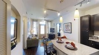 This single-floor residence showcases an open-concept layout that connects the kitchen, dining, and living areas into one cohesive, functional space. The kitchen features a granite-topped breakfast bar with pendant lighting hanging overhead, creating a natural division between the cooking and dining zones while maintaining an open, airy feel. The dining area accommodates a full table and chairs, positioned conveniently between the kitchen and living room. Rich, dark hardwood flooring extends throughout, adding warmth and visual continuity to the space. The living room is generously proportioned, furnished with a comfortable sofa, coffee table, and an area rug layered over the hardwood floors. A ceiling fan is centrally positioned, providing air circulation and additional lighting. Large windows dressed with patterned curtains allow natural light to filter into the living area, enhancing the bright, welcoming ambiance. Track lighting runs along the ceiling, providing flexible, directional illumination. Framed artwork on the walls and thoughtful décor details contribute to the polished, well-finished character of the interior.