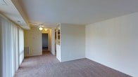 This single-floor space presents a clean, functional layout centered around an open living area finished with wall-to-wall carpet in a neutral tone. The room is defined by smooth, light-colored walls and a standard ceiling height that contributes to a bright, airy feel. A ceiling fan with a built-in light fixture is positioned in an adjoining area, providing both airflow and ambient lighting throughout the connected spaces.

A built-in shelving unit serves as a subtle room divider, separating the main living area from an adjacent space while offering practical storage and display options. The open shelving design keeps the transition between areas visually connected rather than fully enclosed, maintaining a sense of openness throughout.

A hallway extends from the living area, leading to additional rooms beyond. The corridor is carpeted in the same neutral tone, creating a cohesive flow throughout the space. The overall palette of whites and soft beiges gives the interior a neutral, move-in-ready character. The layout is straightforward and practical, making efficient use of the single-floor footprint.