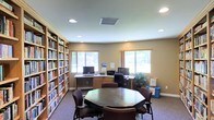 This single-floor space serves as a versatile community or resource room, thoughtfully designed to accommodate both collaborative and individual use. The most striking feature is the floor-to-ceiling built-in bookshelf lining the entire left wall, filled with an extensive collection of books organized across multiple shelves, lending the room a scholarly and welcoming character.

At the center of the room sits an octagonal meeting table surrounded by upholstered chairs, providing a comfortable space for group gatherings or discussions. Along the far wall, an L-shaped built-in desk with warm wood cabinetry houses multiple computer workstations, complete with monitors and a printer, making the space well-suited for productivity and research.

Two windows along the back wall allow ample natural light to fill the room, offering views of greenery outside. Recessed ceiling lights supplement the natural light, ensuring the space remains bright throughout the day. Neutral beige walls and carpeted flooring contribute to a calm, functional ambiance. A potted plant near the workstations adds a touch of warmth to the overall setting.