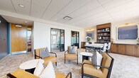 Located at 360 State St in New Haven, Connecticut, this thoughtfully designed lounge space offers a refined and versatile environment suited for both relaxed gatherings and focused meetings. The open floor plan is anchored by a patterned area rug that ties together multiple seating clusters, each furnished with tan leather armchairs accented by houndstooth throw pillows and paired with sleek black side tables in marble and dark wood finishes. A warm-toned built-in wooden bookcase with upper display shelving and lower cabinetry lines one wall, flanking a dining or conference table with upholstered seating — ideal for collaborative work or private meetings. Two large abstract artworks in blue tones add a curated, gallery-like quality to the space. A bold slate-blue accent wall in one seating area provides visual contrast against the otherwise neutral white palette, and is adorned with a trio of framed prints. Floor-to-ceiling windows along the exterior wall invite abundant natural light and offer verdant views of the surrounding New Haven streetscape. A glass-partitioned private room at one end provides an option for more enclosed meetings. Recessed ceiling lighting and a drop tile ceiling complete the polished, professional ambiance throughout.