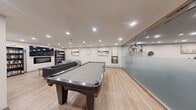 Located at 156-158 Fitch St, this single-floor amenity space is thoughtfully designed to blend fitness and leisure in a clean, modern environment. The fitness area features rubber-floored exercise space equipped with cardio machines including a stationary bike, elliptical, and treadmill, as well as a dumbbell rack and a multi-function cable machine. Wall-mounted mirrors and a flat-screen television enhance the workout experience, while recessed lighting throughout ensures the space is well-lit. Glass partitions with frosted panels elegantly divide the fitness zone from the lounge area, maintaining an open yet purposeful layout. The lounge area is finished with light hardwood-style flooring and features a full-sized billiards table as its centerpiece, complemented by a comfortable sectional sofa and accent chairs. Landscape-inspired artwork and a wall clock add a refined, relaxed touch to the space. A compact kitchenette with white cabinetry, a stainless steel microwave, under-counter beverage refrigerator, and sink provides convenient amenities for residents. The overall design is contemporary and functional, offering a cohesive space for both active and social use.