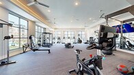 This well-appointed fitness center at 6026 Station Crossing Ave in Charlotte, NC offers a spacious, single-floor layout designed to accommodate a full spectrum of workout routines. The room is flooded with natural light through large, floor-to-ceiling black-framed windows that frame views of the surrounding community and pool area, creating an open and energizing atmosphere. Ceiling fans are distributed throughout the space, complementing the recessed lighting and tray ceiling details that add architectural dimension to the room. Durable patterned carpet covers the floor throughout the main exercise area. The facility is comprehensively equipped with cardio machines including treadmills, ellipticals, and stair climbers by TRUE Fitness, as well as spin bikes and rowing machines. Strength training is well-represented with a full rack of dumbbells, medicine balls, a multi-station cable machine, a power rack, and a punching bag. Functional fitness equipment such as a tire flip station and a sled round out the offerings. Wall-mounted televisions provide entertainment throughout. Full-length mirrors along one wall enhance the sense of space and support form-checking during workouts, making this fitness center a thorough and functional amenity.