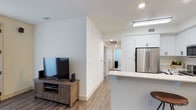 This well-appointed single-floor space showcases a contemporary design with a bright, open layout that seamlessly connects the main living areas. The living area provides a comfortable setting, anchored by a media console and flat-screen television, and is finished with warm-toned luxury vinyl plank flooring that runs throughout the space, adding both durability and a cohesive aesthetic.

Adjacent to the living area, the kitchen is designed for functionality and style. White shaker-style cabinetry extends to the upper walls, offering generous storage, while a stainless steel French door refrigerator serves as a standout appliance. A clean white countertop peninsula defines the boundary between the kitchen and living space, ideal for casual dining or meal preparation. Recessed and flush-mount lighting illuminate the kitchen and hallway efficiently.

A central hallway connects the living and kitchen areas to the private rooms beyond, maintaining a logical and practical flow throughout the unit. The walls are finished in crisp white throughout, enhancing the sense of brightness and openness. Ceiling-mounted HVAC vents ensure consistent climate control. Overall, the space presents a modern, clean, and functional environment suitable for comfortable everyday living.