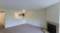 This single-floor residence offers a straightforward and functional layout designed for comfortable everyday living. The living area is finished with plush wall-to-wall carpeting that adds warmth underfoot, while a ceiling fan overhead provides efficient air circulation throughout the space. Smooth, neutral-toned walls and a textured popcorn ceiling contribute to a classic, understated aesthetic.

A defining architectural feature is the pass-through window connecting the living area to the kitchen, promoting an open and communicative flow between the two spaces. The kitchen is equipped with dark wood cabinetry, a white refrigerator, and a range, offering a practical cooking environment. The pass-through counter also serves as a convenient bar-style surface for casual dining or entertaining.

Adjacent to the living area, a tiled platform or hearth is partially visible, suggesting the presence of a fireplace that adds a notable focal point to the room. The overall ambiance is clean, bright, and ready for personalization, making it a practical and livable single-story space well-suited for a variety of lifestyles.