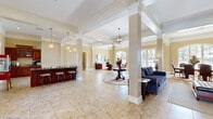 Located at 750 Corder Rd in Warner Robins, Georgia, this single-floor community clubhouse offers a generously sized, open layout designed for both comfort and functionality. The space is anchored by a grand living area distinguished by soaring ceilings, decorative columns with crown molding, and gleaming large-format tile flooring throughout. A striking floor-to-ceiling stone fireplace with a bold wood mantel serves as the room's focal point, complemented by a wall-mounted television and cozy seating arrangements that invite relaxation.

Natural light pours in through multiple arched and French-style windows and doors, creating a bright and welcoming ambiance. A central pedestal table with a floral arrangement adds an elegant touch to the open circulation space. The kitchenette area features rich dark-wood cabinetry with glass-front uppers, a dark granite-topped bar, and counter-height bar stools, all illuminated by pendant lighting. A fun popcorn machine adds a charming, social element to this gathering spot.

A private office area, accessed from the main hall, includes arched windows, polished tile flooring, and refined furnishings, providing a quiet and professional setting. The overall aesthetic blends traditional Southern warmth with upscale finishes, making this a versatile and inviting communal space.