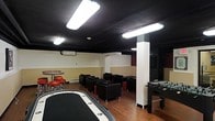 This single-floor entertainment space is purpose-built for social gaming and leisure. The room features a striking black ceiling that creates a moody, club-like atmosphere, contrasted by cream and tan-painted lower walls and warm hardwood flooring throughout. Overhead fluorescent lighting provides bright, even illumination across the space.

The layout is thoughtfully divided into distinct activity zones. A poker table anchors one side of the room, offering a dedicated area for card games. On the opposite end, a foosball table provides additional recreational options. A bar-height table with red stools creates a casual dining or social area near one corner.

The central lounge area is furnished with multiple black leather armchairs and small side tables, arranged to encourage conversation and group gatherings. A structural white column adds an architectural element that subtly separates the space without fully dividing it.

Climate control is provided by a wall-mounted split AC unit, ensuring year-round comfort. The overall design strikes a balance between a relaxed lounge aesthetic and a functional entertainment venue, making it well-suited for private events, game nights, or social club activities.