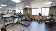 This fitness room is a dedicated exercise space designed to support a variety of cardio-focused workouts. The room is equipped with key cardio machines, including treadmills, an elliptical cross-trainer, and a stationary upright bike, all arranged to allow comfortable use and movement between equipment. The flooring consists of durable dark rubber matting, which provides both safety and stability during workouts. A large wall-to-wall mirror spans one side of the room, enhancing the sense of space and allowing users to monitor their form during exercise. The drop-panel ceiling incorporates recessed fluorescent lighting, ensuring the space is well-lit at all times. Large windows along one wall allow an abundance of natural light to fill the room and offer outward views, contributing to an open and airy atmosphere during workouts. A subtle yellow-green accent stripe runs along the upper portion of the walls, adding a touch of energy and visual interest to the otherwise neutral color palette. A wall-mounted radiator beneath the windows ensures the space remains comfortable year-round. Overall, this fitness room offers a practical, functional environment suited for daily cardiovascular exercise.