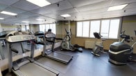 This fitness room is a dedicated exercise space designed to support a variety of cardio-focused workouts. The room is equipped with key cardio machines, including treadmills, an elliptical cross-trainer, and a stationary upright bike, all arranged to allow comfortable use and movement between equipment. The flooring consists of durable dark rubber matting, which provides both safety and stability during workouts. A large wall-to-wall mirror spans one side of the room, enhancing the sense of space and allowing users to monitor their form during exercise. The drop-panel ceiling incorporates recessed fluorescent lighting, ensuring the space is well-lit at all times. Large windows along one wall allow an abundance of natural light to fill the room and offer outward views, contributing to an open and airy atmosphere during workouts. A subtle yellow-green accent stripe runs along the upper portion of the walls, adding a touch of energy and visual interest to the otherwise neutral color palette. A wall-mounted radiator beneath the windows ensures the space remains comfortable year-round. Overall, this fitness room offers a practical, functional environment suited for daily cardiovascular exercise.