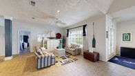 This single-floor space embraces a distinct coastal aesthetic throughout, combining thoughtful design details with functional layouts. The open main area immediately draws attention with its soaring vaulted ceiling and a bold blue spiral staircase serving as a dramatic architectural centerpiece. Tile flooring in a warm stone tone runs consistently throughout, tying the various zones together seamlessly.

The living area is styled with nautical charm, featuring striped sofas, a wicker coffee table, and decorative accents including a model sailboat, a vintage wooden chest, and a decorative oar. A ceiling fan overhead and a large window dressed in navy and patterned curtains contribute to the relaxed, coastal ambiance.

Adjacent to the living area, a dining nook offers seating for several guests around a round table, bathed in natural light from multiple windows also framed by matching navy curtains. Structural columns with a blue-grey finish are present throughout, adding architectural interest while delineating the different zones.

The kitchen features a raised bar with a beadboard-paneled front, granite-style countertops, bar stool seating, dark cabinetry, and a pendant light. Standard appliances include a full-size refrigerator and microwave. Crown molding and textured ceilings are consistent throughout, lending the space a polished, cohesive finish.