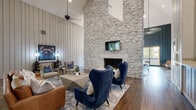 Located at 5901 John F Kennedy Blvd in North Little Rock, Arkansas, this single-floor gathering space makes an immediate impression with its dramatic, soaring ceilings that amplify the sense of openness and grandeur. The undisputed centerpiece of the main lounge is a towering floor-to-ceiling stone fireplace, crafted from light-toned natural rock that extends the full height of the room, anchoring a mounted flat-screen TV above the hearth. Warm hardwood flooring stretches throughout, complementing the vertical shiplap wall paneling rendered in soft neutral tones on one side and a deep charcoal-teal finish on the other, creating a sophisticated contrast. The lounge area is thoughtfully arranged with an eclectic mix of modern furnishings, including plush navy wingback chairs, a cognac leather sofa, and geometric wood coffee tables atop layered area rugs. An elegant sputnik chandelier adds a mid-century modern touch overhead. Large floor-to-ceiling windows and glass doors flood the space with abundant natural light and offer unobstructed views of the lush outdoor greenery beyond. A secondary seating nook with leather lounge chairs sits near the windows, providing an ideal spot for relaxation. A small kitchenette area is also visible, adding everyday convenience. The overall ambiance is warm, inviting, and design-forward.