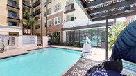 This outdoor amenity space centers around a generously sized, crystal-clear swimming pool that serves as the focal point of a well-appointed courtyard setting. The pool area is enclosed within a modern multi-story residential building, whose facade features a dynamic combination of warm stone cladding, neutral stucco finishes, and private balconies that overlook the courtyard below. Tall palm trees and carefully arranged tropical plantings add a lush, resort-like ambiance to the space. The surrounding pool deck is finished with decorative patterned hardscaping and is furnished with lounge chairs and chaise seating, ideal for relaxing in the sun. An ADA-compliant pool lift is conveniently positioned at the pool's edge, ensuring accessibility for all users. Adjacent to the pool, a glass-enclosed indoor amenity space — likely a fitness center or lounge — provides additional functionality while maintaining visual openness to the outdoor area. Iron fencing defines the perimeter of the pool deck, offering both safety and an unobstructed sightline throughout the courtyard. The overall atmosphere is bright, open, and inviting, with abundant natural light and a well-maintained environment that reflects a high standard of residential amenity design.