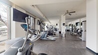 This single-floor fitness center offers a spacious and thoughtfully designed workout environment suited for a wide range of exercise routines. The layout is open and functional, with durable speckled rubber flooring throughout that provides both traction and comfort during workouts.

The cardio zone is lined with multiple elliptical trainers positioned along the large floor-to-ceiling windows, which flood the space with natural light and offer views of the surrounding streetscape. Ceiling fans overhead provide additional air circulation, enhancing comfort during intense sessions.

The strength training area features a variety of cable and resistance machines arranged to allow ample room for movement between stations. A dedicated free weights section includes a full rack of hex dumbbells accompanied by a flat bench, with wall-mounted flat-screen TVs positioned for easy viewing from multiple angles.

Mirrored walls span much of the perimeter, visually expanding the space while allowing users to monitor their form. Structural columns with decorative metal-plated bases are integrated throughout, adding an industrial design detail. The bright white walls and recessed lighting contribute to a clean, modern aesthetic that keeps the environment feeling energetic and motivating.