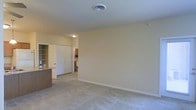 This single-floor space features an open and functional layout designed for comfortable everyday living. The main living area is finished with wall-to-wall carpet in a neutral tone, providing a soft underfoot feel that complements the warm beige walls throughout. A central ceiling vent ensures consistent climate control, while a flush-mounted ceiling light provides overhead illumination.

The living area flows naturally toward an adjoining space, where a partial view reveals built-in shelving that offers practical storage and display options. A paneled door with a bright white finish provides access to an additional room, likely a bedroom or utility area, adding to the overall functionality of the layout.

To the left, a countertop with a dark surface hints at a kitchen or bar area, suggesting a convenient transition between living and dining or food preparation spaces. Cabinetry in a light wood tone is partially visible in the background, contributing to a cohesive and understated interior palette.

Clean white baseboards frame the perimeter of the room, adding a crisp finishing detail. The overall ambiance is calm, neutral, and move-in ready, offering a blank canvas adaptable to a variety of interior styles.