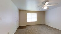This single-story space features a welcoming main room finished with wall-to-wall neutral carpeting that lends a warm, comfortable feel underfoot. Near the entry door, the flooring transitions to durable hard surface material, providing a practical area for arriving and departing. A centrally positioned window allows natural light to fill the room, brightening the neutral-toned walls and contributing to an open, airy atmosphere. A ceiling fan with an integrated light fixture is mounted overhead, offering both lighting and airflow control year-round. The walls are finished in a soft, warm neutral tone that complements the light carpeting, while the textured ceiling adds a subtle visual detail. The entry door is equipped with a standard lock, and a light switch panel is conveniently positioned nearby. The overall layout is straightforward and functional, with clean lines and a simple design that provides a blank canvas adaptable to a variety of furnishing arrangements. The space maintains a modest, practical character throughout.