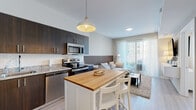 This thoughtfully designed 2-bedroom, 1-bathroom residence offers a contemporary open-concept layout on a single floor. The kitchen is a standout feature, equipped with stainless steel appliances, dark espresso cabinetry, granite countertops, a white subway tile backsplash, and a butcher block island with bar seating beneath a stylish pendant light. Light wood-tone flooring runs throughout, lending visual continuity across the space. The kitchen flows seamlessly into the living room, which is anchored by a slatted wood accent wall and a large sliding glass door that fills the room with natural light and provides access to an outdoor area. The bedrooms are finished in neutral tones with sheer window treatments that allow ample daylight. The primary bedroom features a decorative slatted headboard wall and a window with layered blinds and curtains. The full bathroom is finished with large-format white tile, a granite-topped dark vanity, a combined tub and shower, and a full-width mirror. A hallway closet provides additional storage. The overall design is clean, modern, and cohesive, with a warm neutral palette throughout.