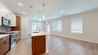 Located at 18000 W 97th St in Lenexa, Kansas, this well-designed two-bedroom, two-bathroom residence offers a thoughtful single-floor layout with modern finishes throughout. The open-concept living area flows seamlessly into the dining space and kitchen, where abundant natural light pours in through multiple windows fitted with blinds. The kitchen is a standout, featuring stainless steel appliances including a refrigerator, range, and microwave, complemented by warm wood-toned cabinetry, white upper cabinets, a tile backsplash, and a central island with a white quartz countertop, undermount sink, and stylish pendant lighting above. Wood-look luxury vinyl plank flooring extends throughout the main living areas, adding warmth and continuity. Both bedrooms are carpeted and include ceiling fans, natural light from windows, and access to generous closets with built-in shelving and wire rack systems. The bathrooms are finished with wood-toned vanities, white countertops, and tiled tub-shower combinations with glass enclosures. A dedicated laundry room houses a full-size top-load washer and dryer alongside overhead wire shelving for added storage. The overall aesthetic is clean and contemporary, with neutral tones creating a bright, inviting atmosphere throughout.