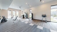 Located at 64–76 E McMillan St in Cincinnati, OH, this well-appointed fitness center offers a comprehensive workout environment across a single, open floor. The space is designed with a clean, contemporary aesthetic, featuring neutral-toned walls, recessed lighting, and ceiling fans that contribute to a comfortable, well-ventilated atmosphere.

The exercise room is divided into distinct functional zones. The cardio area lines the perimeter and is equipped with treadmills and stair climbers, positioned alongside a row of large windows that flood the space with natural light and provide views of the surrounding neighborhood. A dedicated strength training zone houses a multi-station cable machine and additional resistance equipment, complemented by wall-mounted mirrors that visually expand the room and assist with form.

The free weights section features a full rack of dumbbells alongside adjustable benches, making it suitable for a wide range of strength training exercises. Flooring throughout the space combines durable wood-look planks with modular carpet tiles, clearly delineating workout zones while adding a polished, modern finish.

The layout is thoughtfully organized to allow ample space between equipment, promoting ease of movement. A small workstation area near the entrance provides a convenient check-in or administrative point. Overall, this fitness center delivers a professional-grade workout environment with a bright, open ambiance.