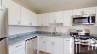 This single-floor space offers a clean, modern aesthetic throughout, with a cohesive neutral color palette of whites and soft beiges. The kitchen serves as a functional centerpiece, featuring white shaker-style cabinetry with brushed nickel hardware, marbled gray stone countertops, and a full suite of stainless steel appliances including a gas range, over-the-range microwave, refrigerator, and dishwasher. Light wood-style flooring runs through the kitchen, lending warmth to the space.

The living areas are bright and open, benefiting from natural light streaming through windows fitted with vertical blinds. Light hardwood-style flooring continues through these common areas, complementing the neutral wall tones and creating an airy, welcoming atmosphere.

The bedrooms are carpeted in a warm beige tone, offering comfort underfoot. Each room includes a window with vertical blinds, providing natural light and privacy. Closet access is available within the bedrooms.

The bathrooms maintain the home's clean, modern style with white shaker vanities, white countertops, round mirrors, brushed nickel fixtures, and light wood-style tile flooring. Grab bars are present in one bathroom, adding a practical accessibility feature throughout this well-appointed space.
