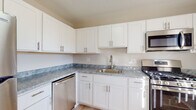 This single-floor space offers a clean, modern aesthetic throughout, with a cohesive neutral color palette of whites and soft beiges. The kitchen serves as a functional centerpiece, featuring white shaker-style cabinetry with brushed nickel hardware, marbled gray stone countertops, and a full suite of stainless steel appliances including a gas range, over-the-range microwave, refrigerator, and dishwasher. Light wood-style flooring runs through the kitchen, lending warmth to the space.

The living areas are bright and open, benefiting from natural light streaming through windows fitted with vertical blinds. Light hardwood-style flooring continues through these common areas, complementing the neutral wall tones and creating an airy, welcoming atmosphere.

The bedrooms are carpeted in a warm beige tone, offering comfort underfoot. Each room includes a window with vertical blinds, providing natural light and privacy. Closet access is available within the bedrooms.

The bathrooms maintain the home's clean, modern style with white shaker vanities, white countertops, round mirrors, brushed nickel fixtures, and light wood-style tile flooring. Grab bars are present in one bathroom, adding a practical accessibility feature throughout this well-appointed space.