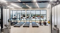 This fitness center offers a thoughtfully designed, single-level space that balances functionality with modern aesthetics. The open layout accommodates a comprehensive range of equipment, including treadmills, stationary bikes, cable machines, weight benches, free weights, and multi-use strength stations, providing a well-rounded workout environment.

Architecturally, the space features a striking dark ceiling with linear LED lighting that runs the length of the room, creating a sleek, polished atmosphere. Light-toned carpet flooring contrasts cleanly with the dark ceiling, contributing to a bright and inviting feel. One wall is adorned with a distinctive illustrated mural, adding a creative, artistic element to the otherwise modern interior.

Floor-to-ceiling windows line one side of the space, flooding the interior with natural light while offering views of an adjacent outdoor area. Glass-paneled entry doors maintain a sense of openness, visually connecting the fitness center to neighboring spaces. Mirrors are strategically placed to enhance the sense of spaciousness throughout.

The thoughtful equipment arrangement allows for clear circulation paths, making the space practical and accessible. Overall, the fitness center presents a contemporary, well-appointed environment suited for a full range of training activities.