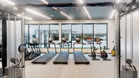This fitness center offers a thoughtfully designed, single-level space that balances functionality with modern aesthetics. The open layout accommodates a comprehensive range of equipment, including treadmills, stationary bikes, cable machines, weight benches, free weights, and multi-use strength stations, providing a well-rounded workout environment.

Architecturally, the space features a striking dark ceiling with linear LED lighting that runs the length of the room, creating a sleek, polished atmosphere. Light-toned carpet flooring contrasts cleanly with the dark ceiling, contributing to a bright and inviting feel. One wall is adorned with a distinctive illustrated mural, adding a creative, artistic element to the otherwise modern interior.

Floor-to-ceiling windows line one side of the space, flooding the interior with natural light while offering views of an adjacent outdoor area. Glass-paneled entry doors maintain a sense of openness, visually connecting the fitness center to neighboring spaces. Mirrors are strategically placed to enhance the sense of spaciousness throughout.

The thoughtful equipment arrangement allows for clear circulation paths, making the space practical and accessible. Overall, the fitness center presents a contemporary, well-appointed environment suited for a full range of training activities.