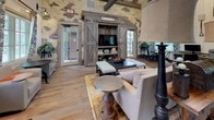 This single-floor community space showcases a striking blend of rustic and industrial design, characterized by exposed natural stone walls, reclaimed wood beams, and wide-plank hardwood flooring throughout. The soaring vaulted ceilings with visible timber framing lend an open, airy quality to the entire space, while industrial-style barn lighting fixtures add warmth and character.

The main lounge area serves as a central gathering point, anchored by a reclaimed wood entertainment unit with sliding barn doors framing a mounted television. Comfortable seating arrangements are complemented by layered rugs and an eclectic mix of furnishings that balance rustic charm with modern comfort. Large windows and glass doors flood the space with natural light and provide views of an outdoor pool area.

A fully equipped kitchen features dark wood cabinetry, stainless steel appliances including double ovens and a refrigerator, and a substantial island with bar seating — ideal for communal use and events. Adjacent to the kitchen, a dining area with a reclaimed wood farm table and black metal chairs creates an inviting space for casual meals or gatherings.

A dedicated business center offers workstations and computing equipment, framed by the same floral-curtained windows and shiplap walls found throughout. Decorative touches such as vintage farm tools, potted ferns, and botanical drapery tie the space together with a cohesive, welcoming aesthetic.