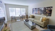 This thoughtfully designed single-floor space offers a seamless blend of modern style and comfortable living. The open living and dining area is bathed in natural light streaming through large windows fitted with horizontal blinds, creating a bright and airy atmosphere throughout the day. Light hardwood flooring anchors the space, adding warmth and continuity across the room.

The living area is centered around a tufted sage-green sofa accented with olive and cream throw pillows, paired with a striking mirrored coffee table that adds a contemporary, reflective element to the room. A large-scale artwork hangs prominently on the wall, serving as a focal point that brings visual depth and character to the space.

The dining nook sits comfortably beside the expansive windows, featuring a round pedestal table paired with woven accent chairs, creating an intimate setting for meals. A tall decorative planter with sculptural branches adds an organic, natural touch to the corner, complementing the neutral color palette of whites, greens, and warm tones found throughout. The overall ambiance is clean, calm, and inviting — well-suited for both everyday living and casual entertaining.