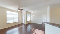 This single-floor home offers a welcoming and well-lit interior defined by clean lines and a thoughtfully open layout. The main living and dining area is a standout feature, showcasing rich, dark hardwood flooring that runs throughout and reflects the abundant natural light streaming in through multiple large windows. Vaulted ceilings lend a sense of spaciousness and airiness to the room, while crown molding along the walls adds a refined architectural touch. A ceiling fan with a light fixture provides both comfort and function, complementing a decorative pendant light that hangs above the dining area, adding a warm, inviting glow. The neutral, light-toned walls create a versatile backdrop suited to a variety of interior styles. A textured ceiling adds subtle visual interest overhead. The open flow between the living and dining spaces connects naturally to additional rooms, visible through doorways that hint at further comfortable living areas beyond. A partial view of a countertop at the edge of the frame suggests a nearby kitchen. The overall ambiance is bright, airy, and move-in ready, combining practical design elements with classic details that give the home a polished, livable character.
