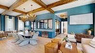 Located at 4711 Azalea Landing Dr in Wilmington, NC, this thoughtfully designed single-floor amenity space blends coastal charm with sophisticated, contemporary interiors. The open-concept living and dining area serves as the heart of the space, showcasing soaring ceilings adorned with rustic exposed wood beams, light hardwood flooring, and bold teal accent walls that set a vibrant yet refined tone. Floor-to-ceiling black-framed windows flood the area with natural light and offer views of the surrounding landscape, while decorative woven chandeliers add warmth and texture overhead. A built-in wet bar with teal cabinetry and granite countertops provides a functional and stylish centerpiece between the living and dining zones. The formal dining room features a striking live-edge wood conference table beneath a sculptural cluster of woven pendant lights, complemented by tall windows and a wall-mounted display screen — ideal for gatherings or presentations. A secondary dining area offers a long communal table with bar-height seating, a tiled accent wall in deep teal mosaic, and additional cabinetry with a sink. The private office is an intimate, glass-enclosed workspace with bold geometric wallcovering, soft sconce lighting, and an outdoor view. Throughout, the design balances coastal warmth with modern functionality.