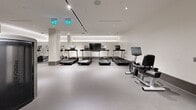 This expansive, single-floor fitness facility offers a comprehensive workout environment thoughtfully organized into distinct training zones. The space is defined by its clean, modern aesthetic — featuring smooth neutral-toned flooring throughout, crisp white walls, and a sophisticated combination of recessed lighting and track lighting that ensures the entire facility is consistently and evenly illuminated. Continuous cove lighting along ceiling perimeters adds a polished architectural accent while contributing to the bright, energizing ambiance.

The cardio zone is equipped with a row of treadmills, each fitted with individual display screens, alongside recumbent bikes and additional cardio machines. A dedicated strength training area houses an extensive selection of dumbbells on tiered racks, cable machines, resistance equipment, and free weight benches. Full-length mirrors line multiple walls throughout the facility, amplifying the sense of space and allowing users to monitor their form.

A separate, more enclosed studio space offers an open floor plan ideal for group classes or individual training, complete with mirrored walls and track lighting. Warm wood-paneled accents and glass-paneled doors introduce natural material contrasts against the otherwise minimalist palette. The overall design balances functionality with a refined, upscale aesthetic, creating a focused and professional environment suited for a wide range of fitness activities.