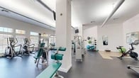 Located at 4440 SW Archer Rd in Gainesville, Florida, this fitness center offers a generously sized, single-floor exercise room designed to support a comprehensive workout experience. The space features high ceilings and an abundance of natural light streaming through large, multi-pane windows fitted with horizontal blinds, creating a bright and energizing atmosphere. White walls and structural columns contribute to the clean, open aesthetic throughout.

The exercise room is thoroughly equipped with a wide range of fitness equipment. Cardio options include multiple treadmills, elliptical trainers, and a stationary cycling bike. A robust selection of Cybex strength machines targets various muscle groups, complemented by a dumbbell rack and medicine ball storage for free-weight and functional training. Large wall-mounted mirrors expand the sense of space and assist with form during workouts. A dedicated stretching and floor exercise area features a wood-toned mat surface, providing a distinct zone for flexibility work and cool-downs. Wall-mounted televisions are positioned throughout the room for entertainment during workouts. The durable rubber flooring offers both safety and comfort underfoot. The overall layout is thoughtfully organized, allowing for smooth circulation between workout zones while accommodating multiple users simultaneously.