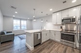 This contemporary single-floor unit offers a well-designed open-concept layout that seamlessly connects the kitchen and living area. The kitchen is a standout feature, showcasing white shaker-style cabinetry, a quartz waterfall peninsula with an undermount sink, and a full suite of stainless steel appliances including a range, built-in microwave, and refrigerator. A stainless steel backsplash adds an industrial touch, while a trio of polished chrome pendant lights hang above the peninsula, providing both task lighting and visual interest. The flooring throughout is a warm-toned wood-look plank, lending a cohesive, modern aesthetic to the entire unit. The living area offers natural light through large windows with urban views of the surrounding neighborhood. The bedroom is calm and airy, featuring soft gray walls, wood-plank flooring, and multiple windows that frame expansive city views, filling the room with natural light. A secondary room with a window provides flexible space suitable for use as a lounge or additional bedroom. The bathroom is outfitted with a clean white tub-shower combination and chrome fixtures. Recessed lighting throughout the unit ensures consistent illumination, while roller shades on the windows provide privacy and light control.