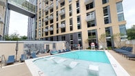 This contemporary multi-story residential building presents a well-designed environment that balances modern aesthetics with functional amenities. The outdoor amenity area serves as a central highlight, featuring a resort-style swimming pool with built-in in-water lounge chairs and a surrounding deck furnished with blue chaise lounges and cushioned seating. The pool area is framed by the building's tall facade, finished in warm beige tones with rows of large windows and private balconies extending from upper floors, creating an enclosed courtyard feel that adds a sense of privacy and scale.

The building's interior common corridors are thoughtfully designed with a consistent, contemporary palette. Wide hallways are lined with smooth, light-gray walls accented by bold teal vertical trim elements that lend a modern edge to the design. Recessed ceiling lighting runs the length of each corridor, ensuring even illumination throughout. The floors are covered in a patterned carpet in shades of gray, teal, and charcoal, reinforcing the building's cohesive color scheme. Unit entry doors are finished in a clean gray tone and feature individual unit numbers accompanied by decorative tile art, providing each residence with a distinct, personalized touch at its entrance.